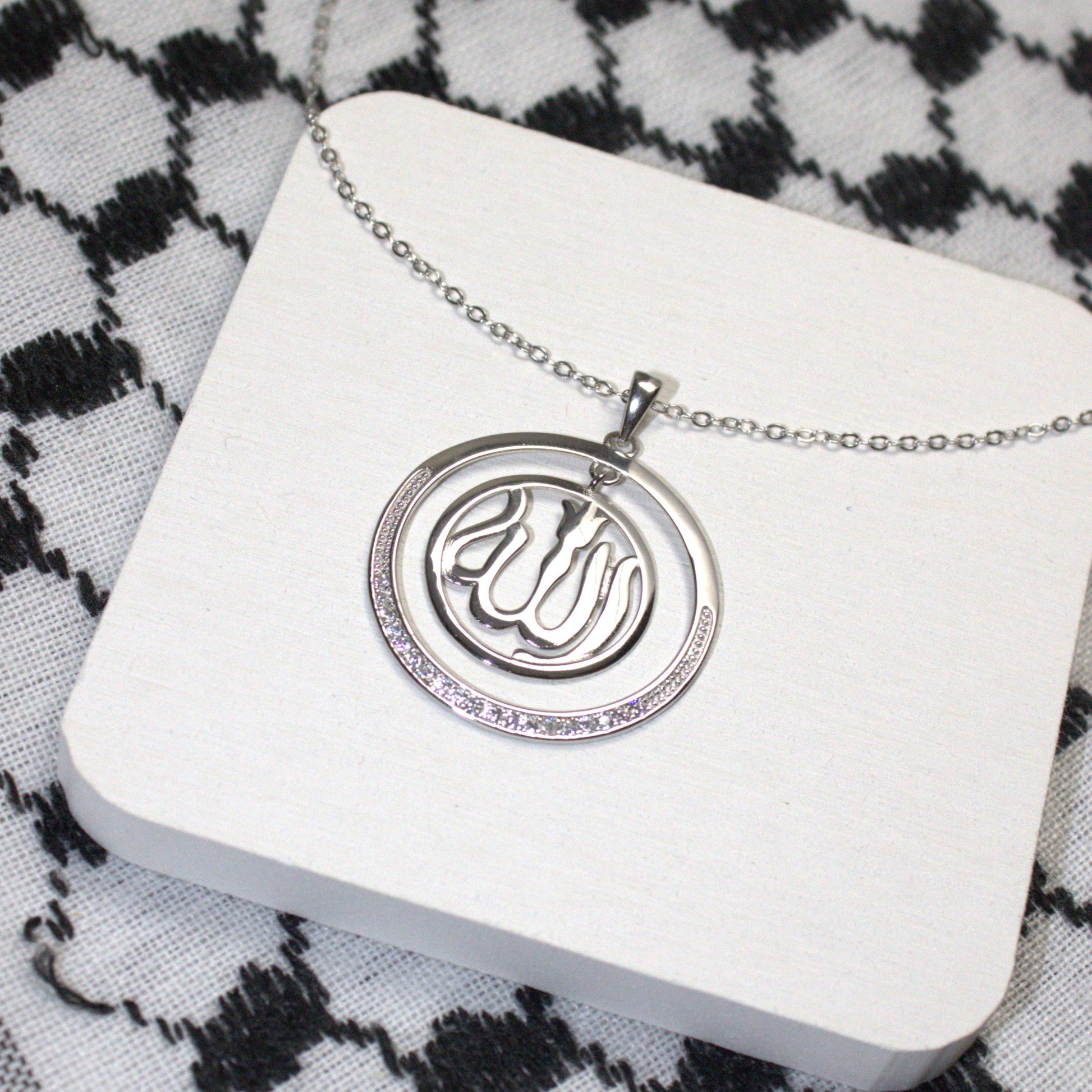 Silver necklace with a pendant on a white card against a black and white keffiyeh background


